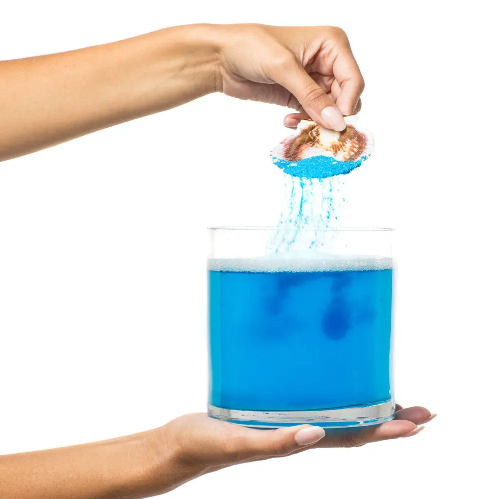 Blue bath salts sprinkling into a glass of water, shown being poured by hands